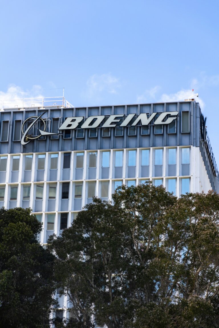 Exterior view of the Boeing corporate building with the Boeing logo prominently displayed on the upper part of the structure, framed by trees in the foreground against a clear blue sky.