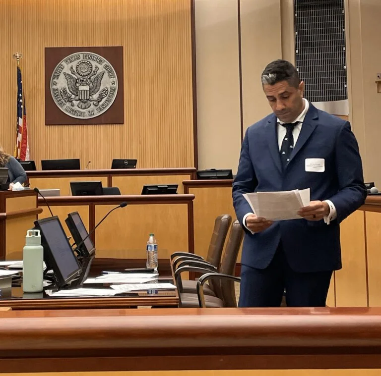 Attorney Mo Houmoudi stands in a courtroom, reading from a set of papers, wearing a navy suit and a determined expression, with the United States District Court emblem displayed on the wall behind him